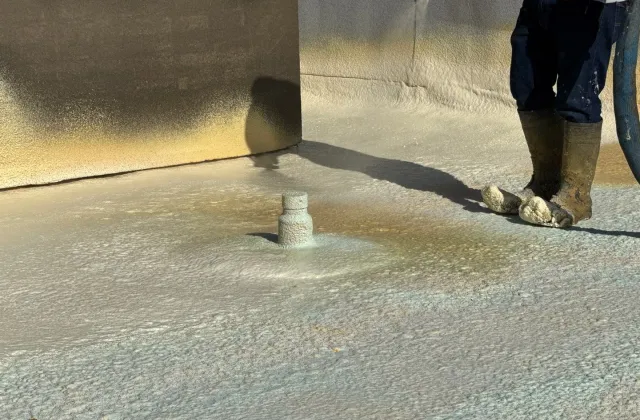 Spray polyurethane foam going down over a roof deck and up a parapet wall, the applicator's boots at the edge of the fresh foam.