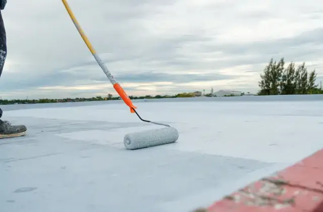 A CES Roofing crew member walking white silicone coating out along a parapet with a roller on a long pole.