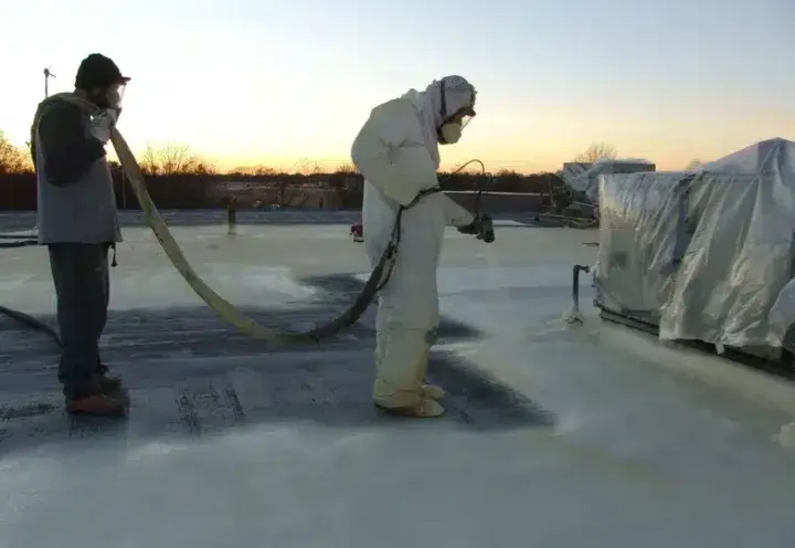Two CES Roofing crew members spraying polyurethane foam across a flat commercial roof in low winter light, one running the hose, one on the gun.