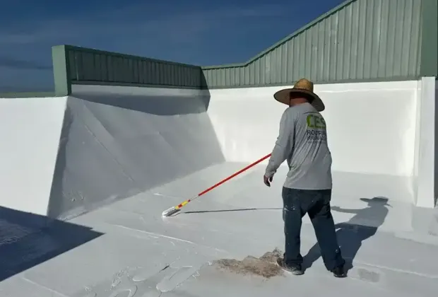 A CES Roofing crew member rolling white coating up a parapet wall on a flat roof under a clear sky.
