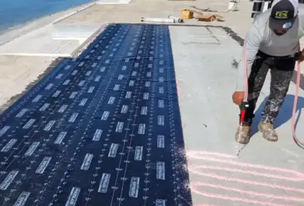 A CES Roofing crew member torching a roll of modified bitumen membrane down onto a concrete deck beside the water.